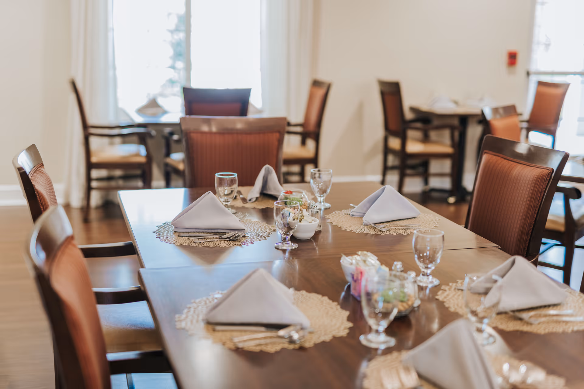 A dining room with wooden tables and chairs set with folded white napkins, glassware, and small bowls of condiments. The room has large windows allowing natural light to brighten the space.