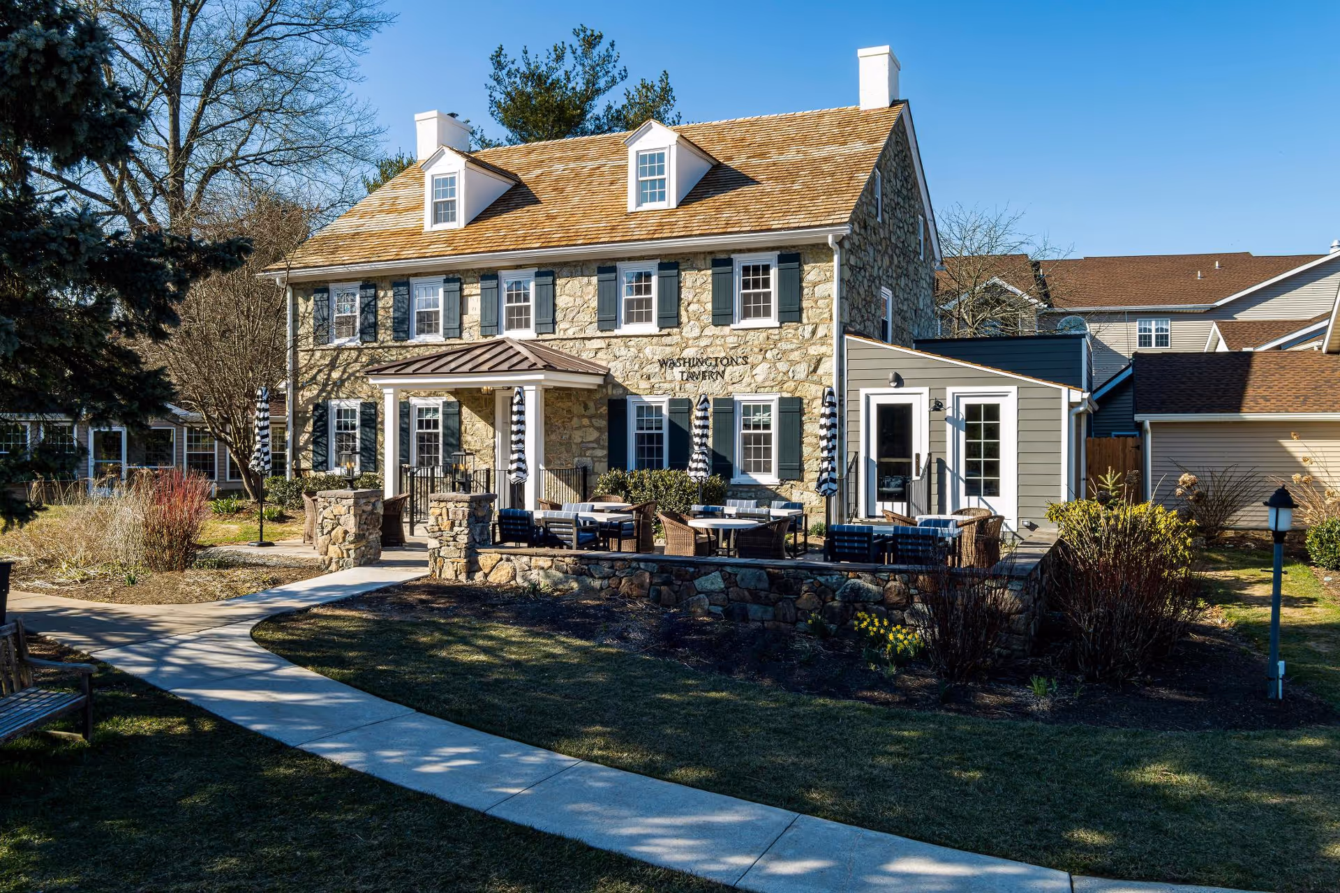 Stone and wood building with a porch and outdoor seating area featuring tables, chairs, and umbrellas. The building is surrounded by a landscaped garden with trees, bushes, and a paved walkway leading to the entrance. The sky is clear and blue.