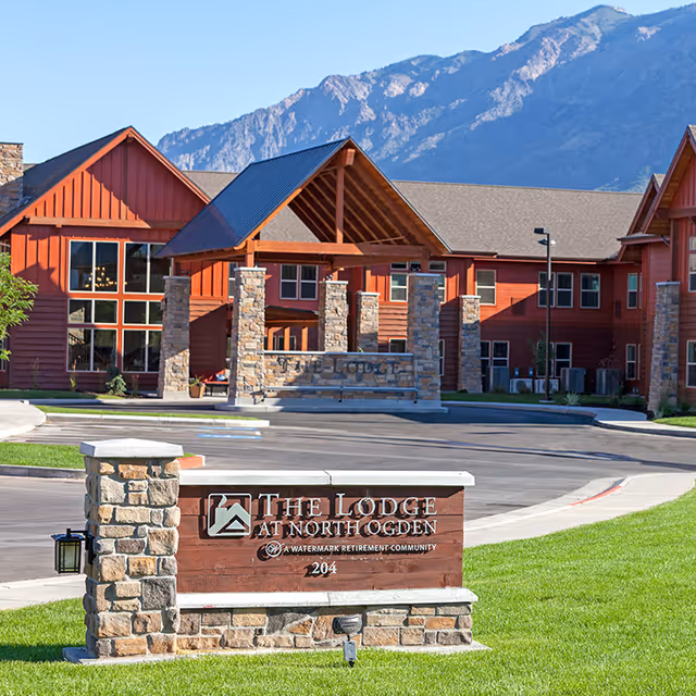 Exterior view of The Lodge at North Ogden, a senior living facility with a rustic design featuring red wooden buildings, stone pillars, and a covered entrance. Mountains are visible in the background under a clear blue sky.