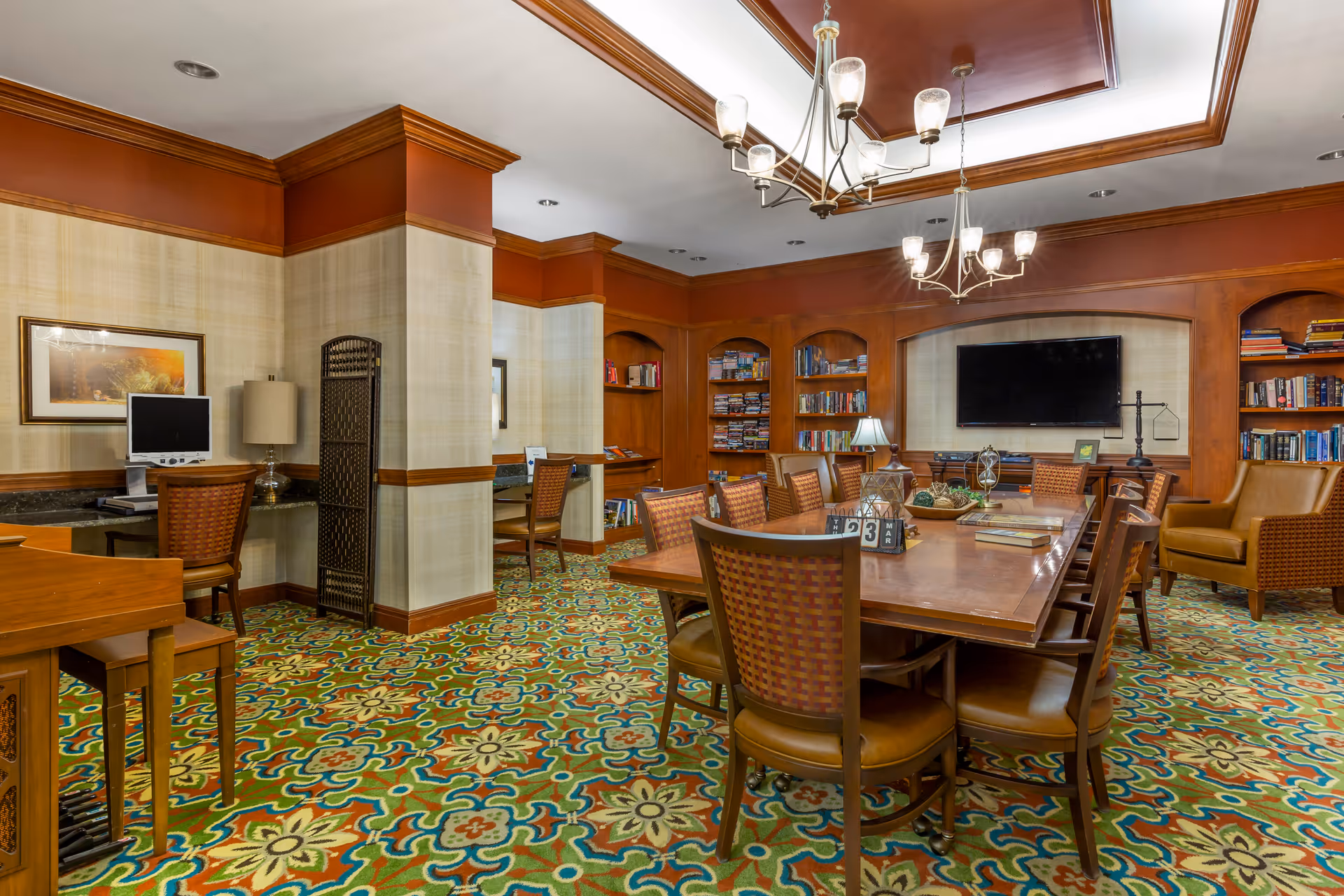 Community room with a large wooden table and chairs, bookshelves, a wall-mounted TV, and computer stations.