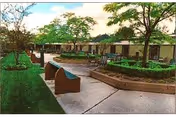 Outdoor courtyard area with paved walkways, benches, and landscaped greenery including trees and bushes, adjacent to a single-story building under a partly cloudy sky.