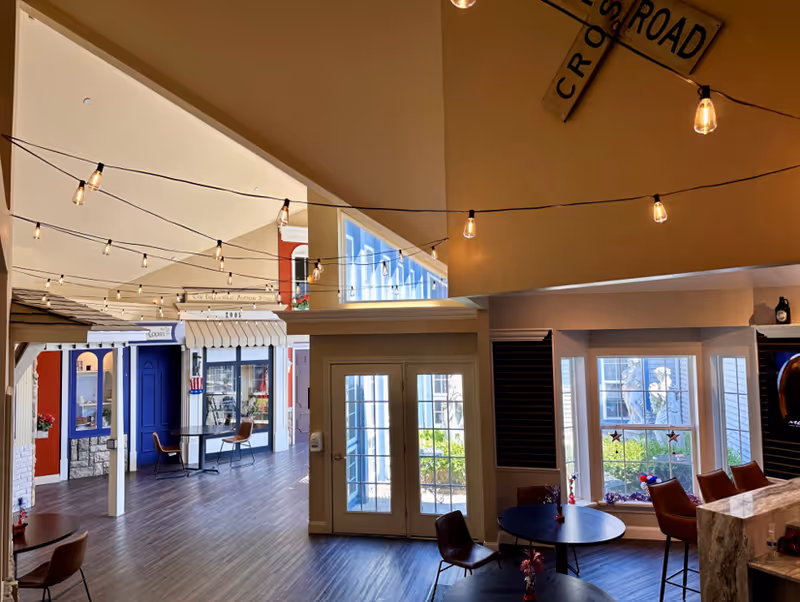 Indoor common area designed like a small town street with storefront facades, string lights hanging from the ceiling, tables and chairs for seating, and large windows letting in natural light.
