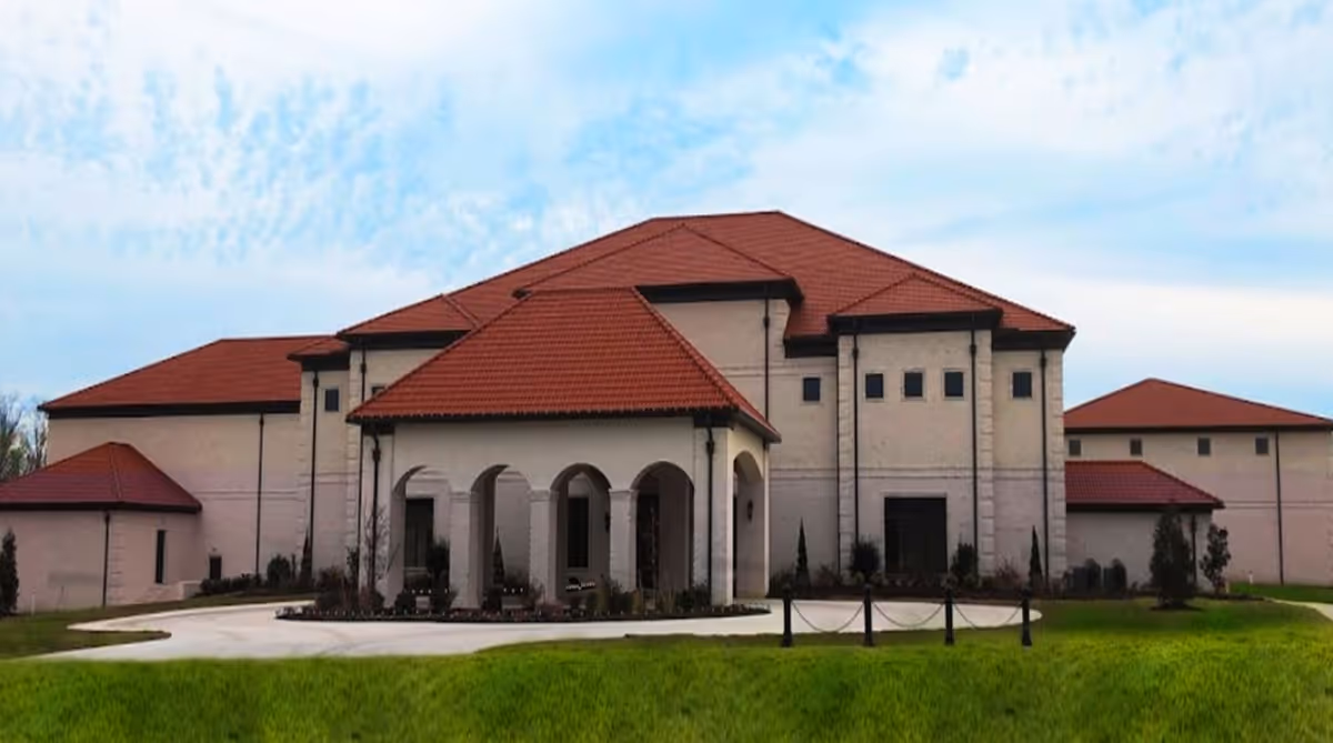 Exterior view of a large building with beige walls and red tiled roofs, featuring an arched entrance and surrounded by green grass and a paved driveway.
