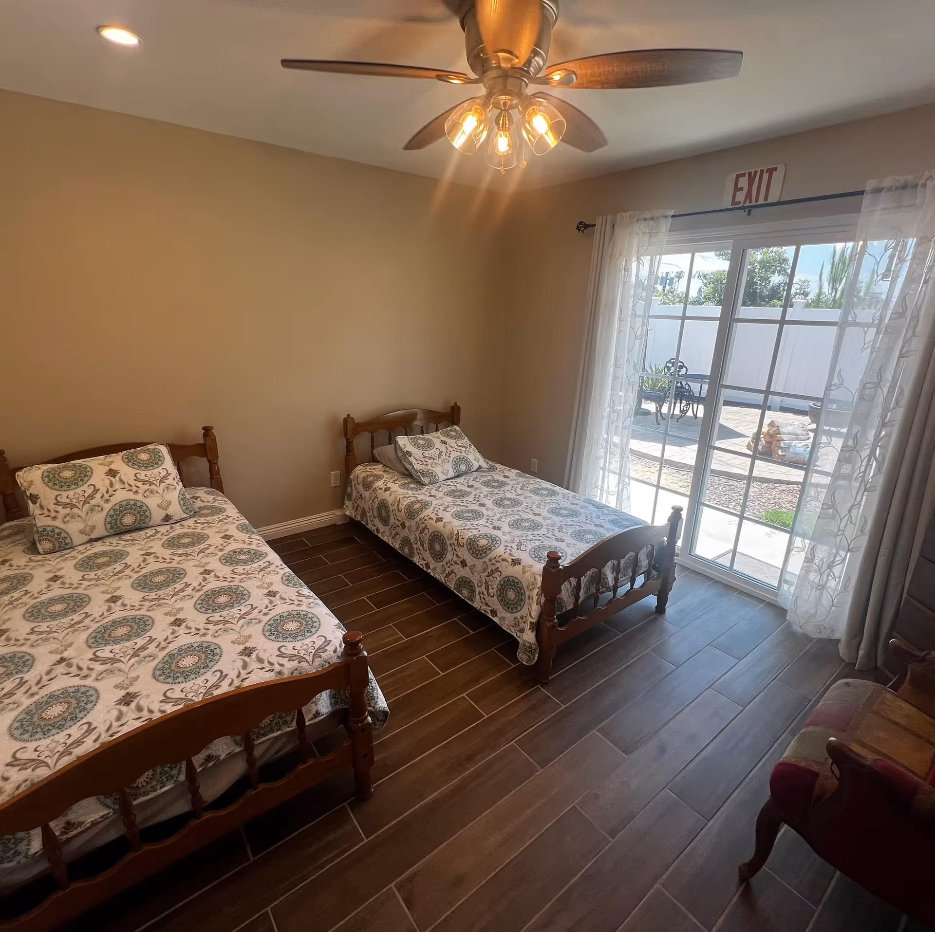Bright bedroom with two wooden twin beds, patterned bedding, a ceiling fan, and a sliding glass door opening to an outdoor patio.