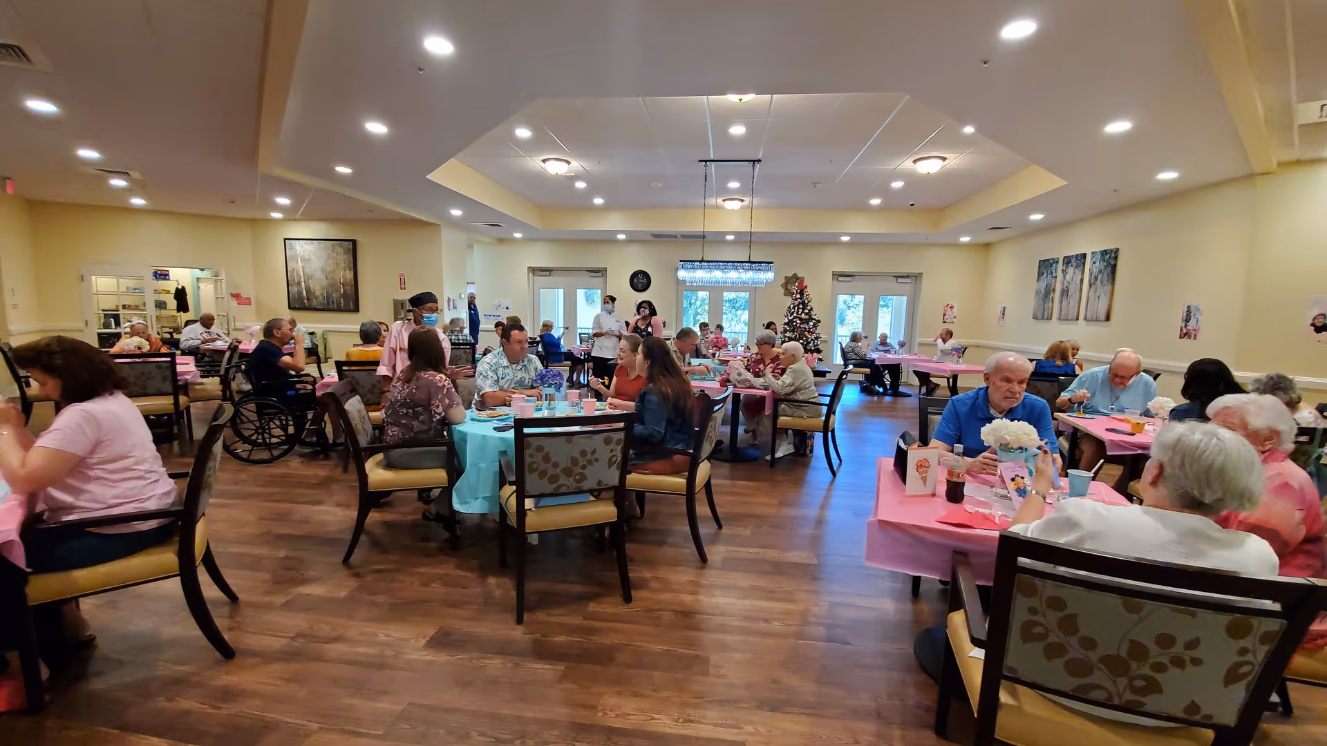A large dining room in a senior living facility with multiple tables covered in pink and blue tablecloths. Elderly residents and staff are seated and standing around the tables, engaging in conversation and eating. The room has wooden floors, cream-colored walls, ceiling lights, and a decorated Christmas tree near the back doors.