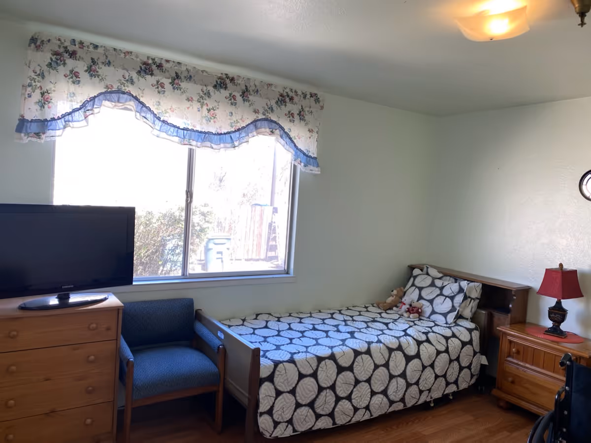 A small bedroom with a single bed covered in a black and white leaf-patterned bedspread. There are two stuffed animals on the bed. Next to the bed is a wooden nightstand with a red lamp. A wooden dresser with a flat-screen TV on top is positioned near a window with floral curtains. A blue upholstered chair is placed between the dresser and the bed. The room has light-colored walls and wooden flooring.