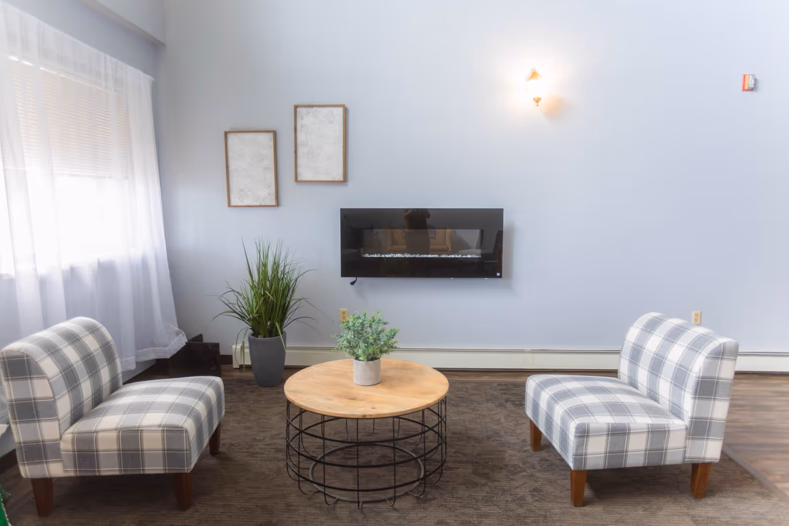 A bright seating area with two plaid chairs facing a round wooden coffee table, a wall-mounted electric fireplace, and potted plants.