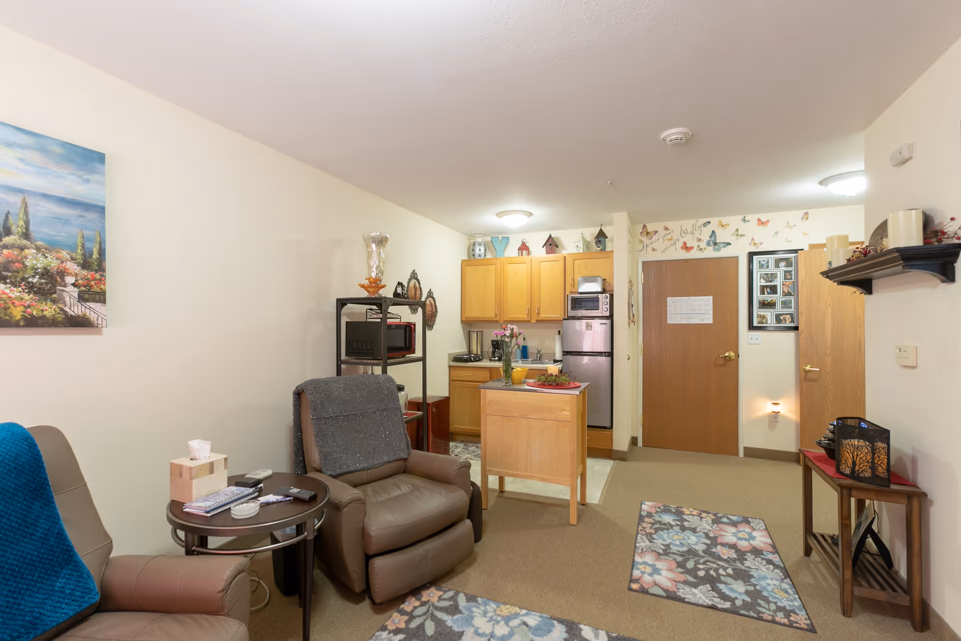 Cozy studio-style living area with recliner chairs, a small kitchen with wooden cabinets and island, and a decorated entry door.