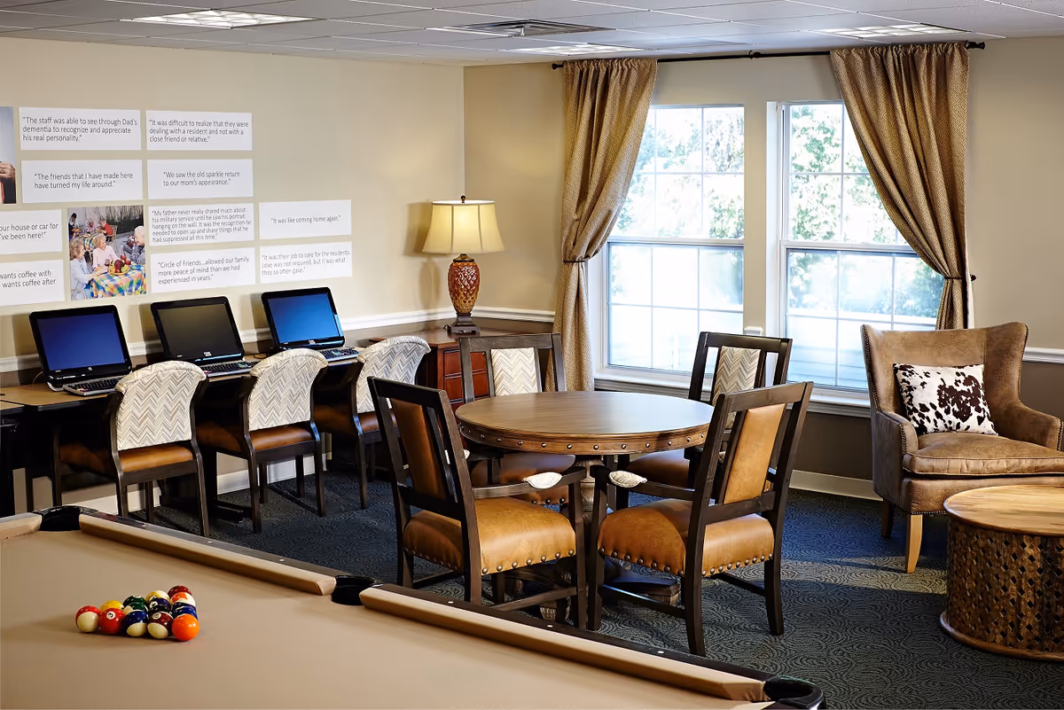 A cozy senior living common area with a pool table in the foreground, a round wooden table with four chairs in the center, a comfortable armchair with a patterned pillow near a window with beige curtains, and a row of four computers on a desk against the wall. The wall has several framed quotes and photos. A table lamp is placed on a small wooden cabinet in the corner.