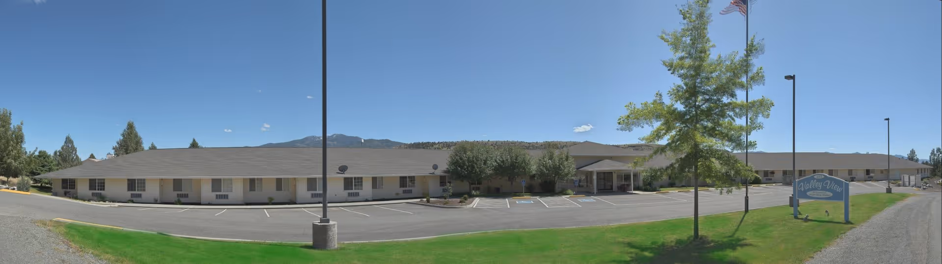 Wide panoramic view of a single-story senior living facility building named Sapphire at Valley View, with a parking lot in front, a green lawn, a flagpole with an American flag, and a sign that reads Valley View. The sky is clear and blue with a few small clouds.