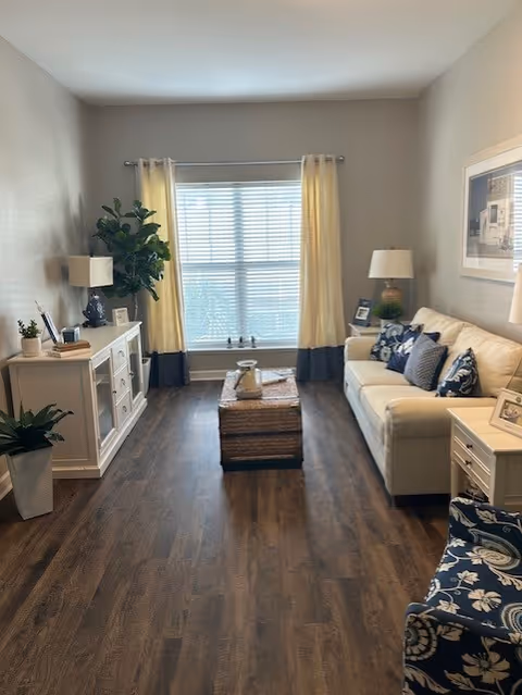 A cozy living room with a white sofa adorned with patterned cushions, a wicker coffee table in the center, a white sideboard with a lamp and decorative items on the left, a large window with light yellow curtains, and a patterned armchair on the right.
