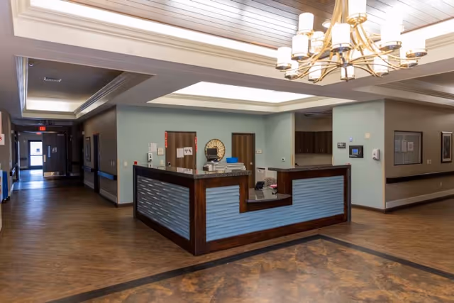Interior view of a reception desk area in a senior living facility with wood flooring, light green walls, and a modern chandelier hanging from the ceiling. The reception desk has a blue panel front with dark wood trim. Hallways extend from the reception area with doors and framed pictures on the walls.