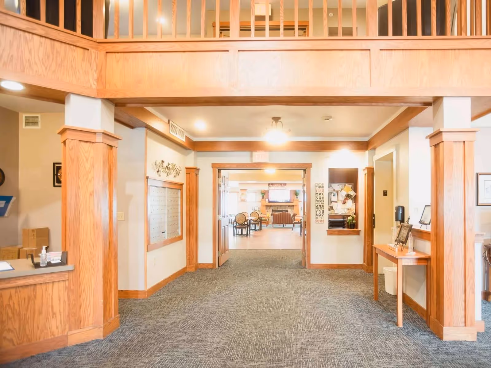Interior view of a senior living facility hallway with wooden pillars and trim, carpeted floor, and a reception desk on the left. The hallway leads to a common area with chairs and a fireplace in the background.