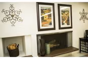 A cozy interior space featuring a white brick fireplace with a wooden mantel. On the mantel are decorative vases and a small stack of books. Above the fireplace hang two framed landscape paintings. On either side of the paintings are symmetrical metal wall decorations. To the left of the fireplace is a basket filled with firewood, and to the right is a black cabinet with a decorative item on top.