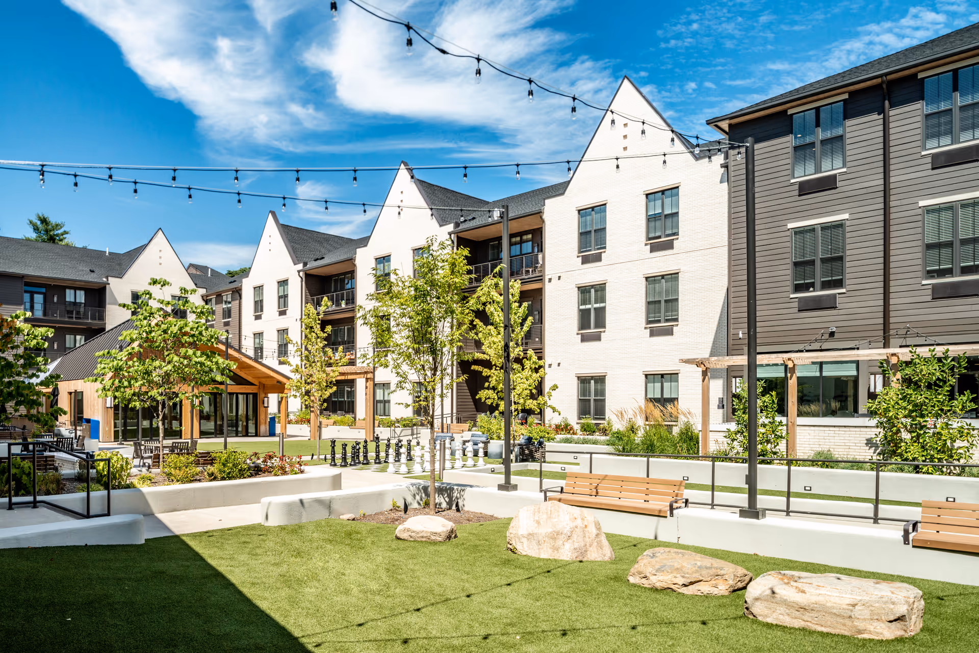 Outdoor courtyard area of a senior living facility with green artificial turf, large rocks, wooden benches, small trees, string lights overhead, and a large chess set. The background shows a multi-story building with white and gray exterior walls and multiple windows under a blue sky with some clouds.