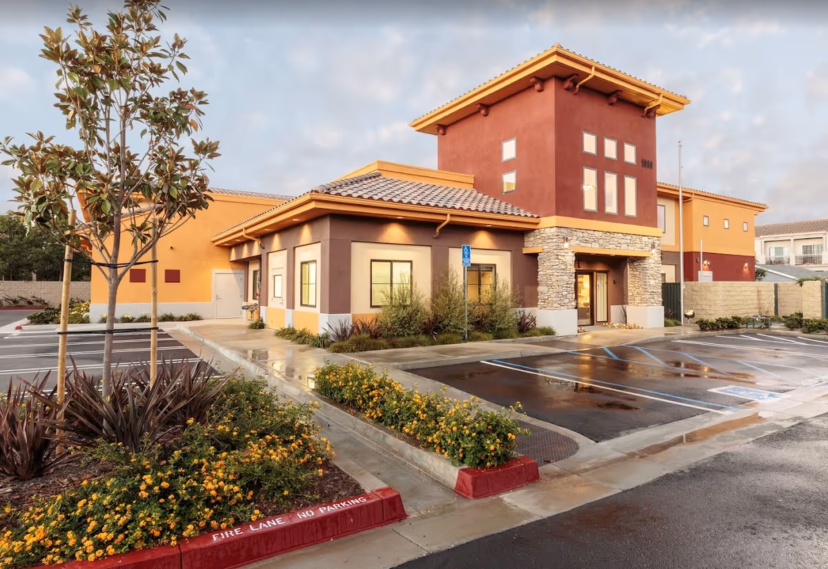Exterior front of a modern memory care building with tiled roofs, stone accents, landscaped flowerbeds and a wet parking lot with handicapped spaces.