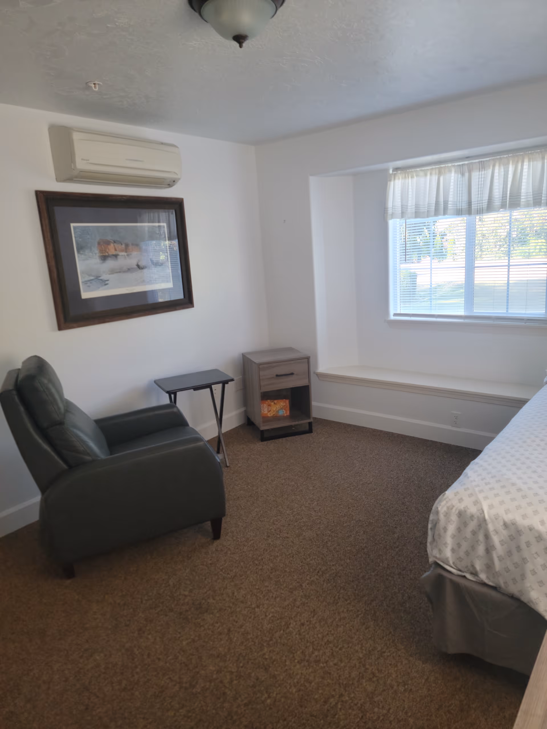 Small bedroom with a green armchair, small side tables, a window seat and part of a bed visible.