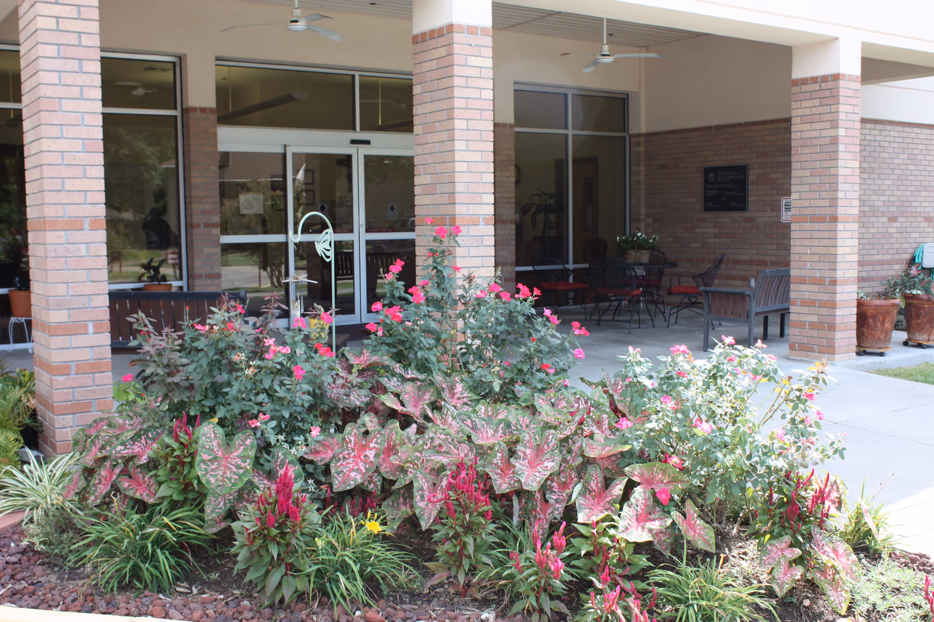 Entrance area of a senior living facility with brick pillars and glass doors. In front, there is a garden bed with various flowering plants including pink flowers and green foliage. Behind the garden, there is a covered patio with outdoor seating including chairs and benches.