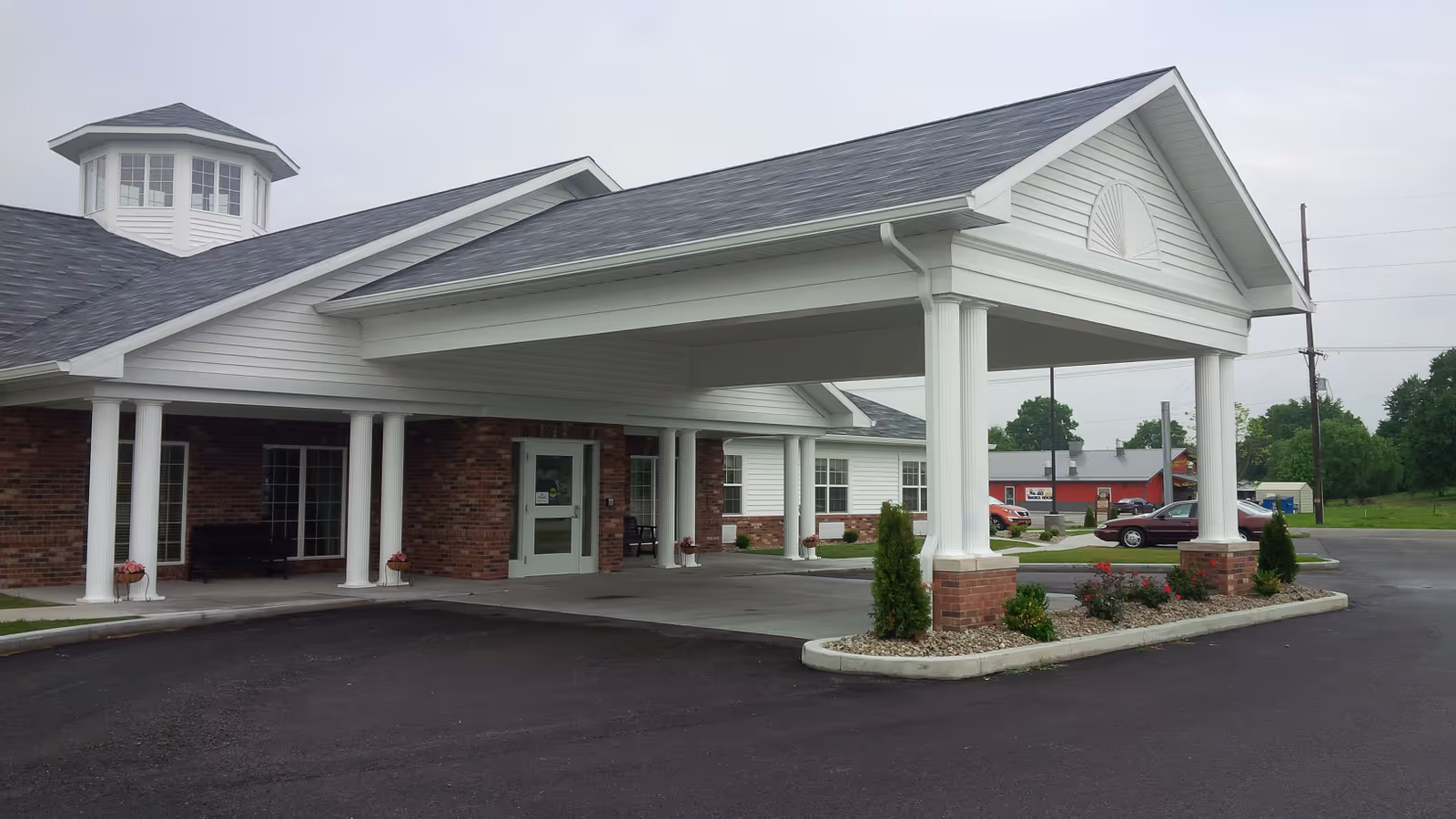 Covered entrance and porte-cochere of a single-story brick and white-sided care building with columns and a driveway.