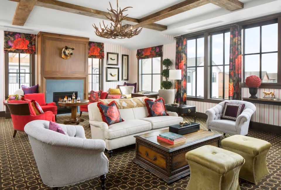 A bright and spacious living room with large windows, floral curtains, and a patterned carpet. The room features a white sofa with red floral pillows, two armchairs, two green ottomans, a wooden coffee table with books, and a fireplace with a mounted animal head above it. There is also a chandelier made of antlers hanging from the ceiling and various decorative items including framed artwork and plants.