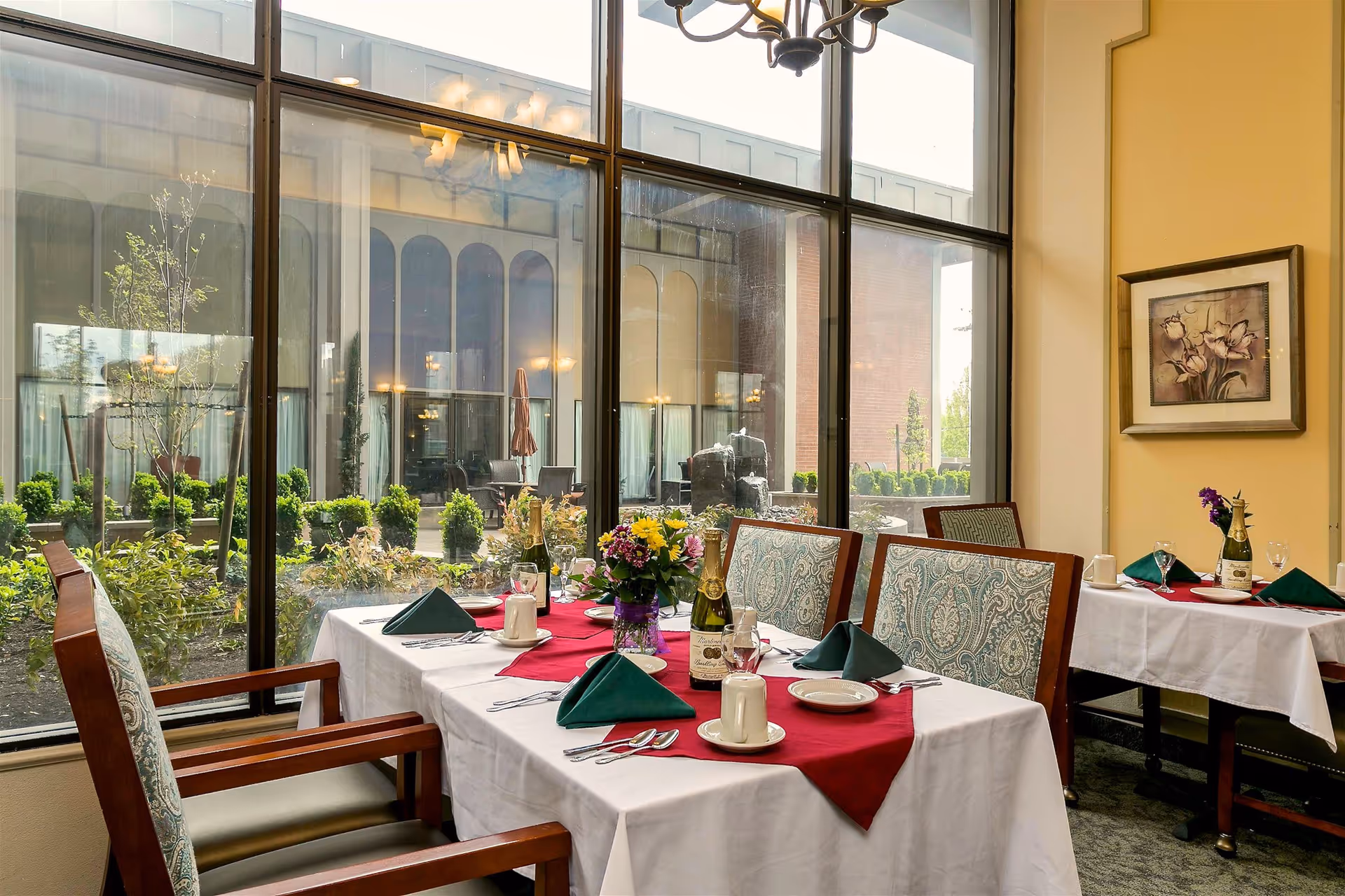 Sunlit dining room with tables set with red runners, green napkins, floral centerpieces, and large windows overlooking a courtyard.