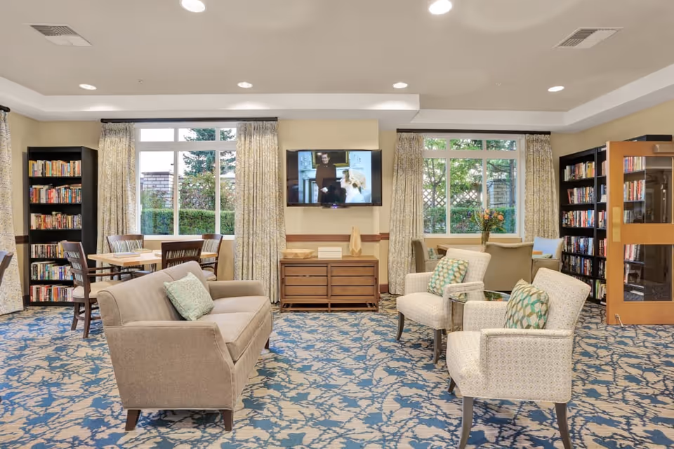 A cozy and well-lit common area with a beige sofa and two patterned armchairs arranged around a small glass-top table. Behind the seating area, there are two large windows with curtains, letting in natural light and showing greenery outside. A flat-screen TV is mounted on the wall above a wooden cabinet. Bookshelves filled with books flank the windows on both sides. There are also tables and chairs near the windows for reading or socializing.