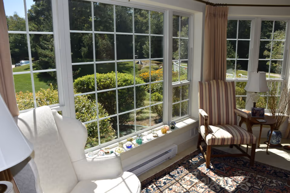 A bright sitting area with large windows overlooking a green garden. The room features a white upholstered armchair, a striped armchair, a small wooden side table with a lamp and framed photo, and a decorative rug on the floor. Various small decorative glass items are displayed on the windowsill.