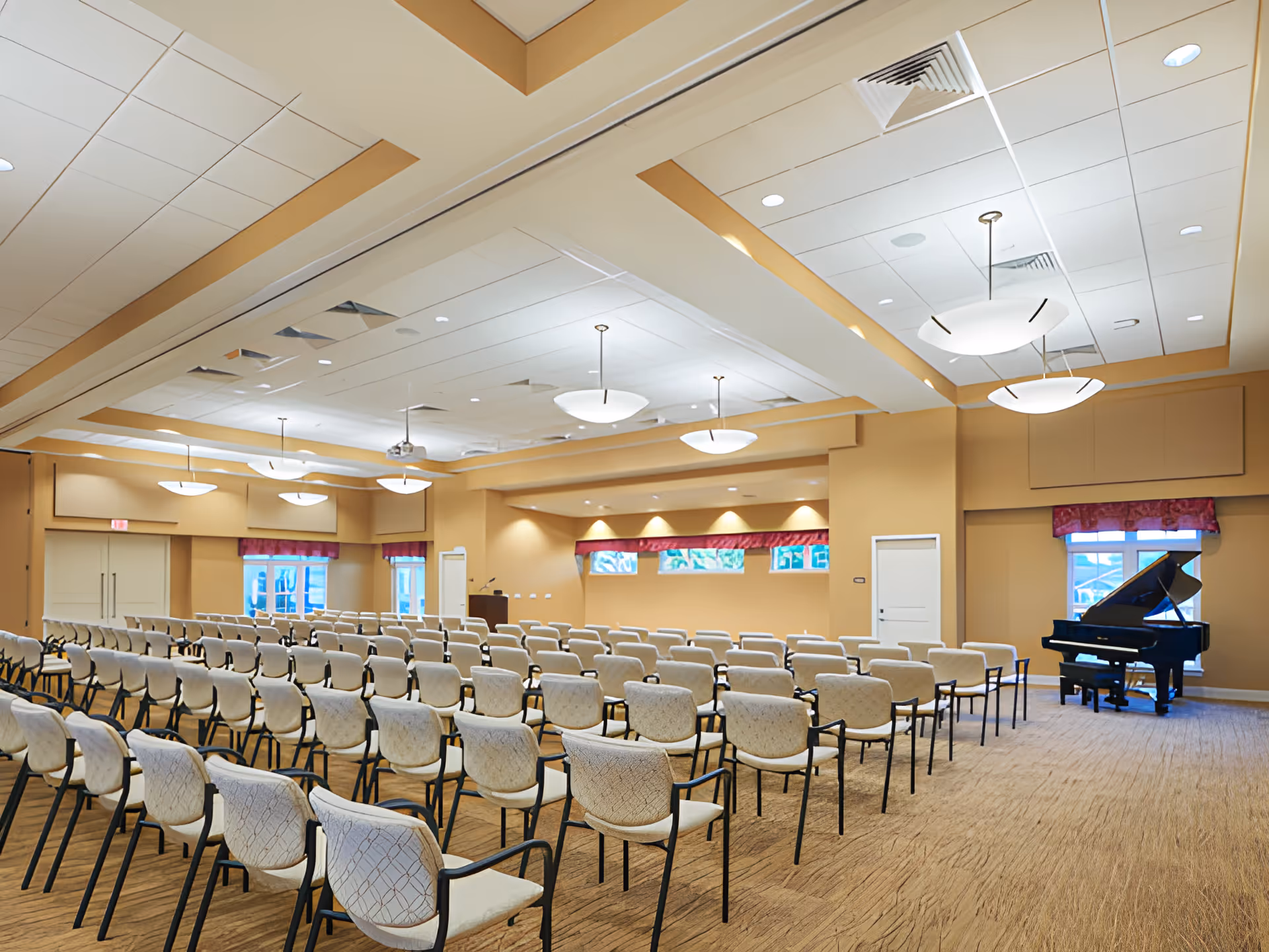 A large meeting or event room with rows of beige cushioned chairs arranged facing a podium and a grand piano near the windows. The room has beige walls, carpeted floor, and ceiling lights with a modern design.