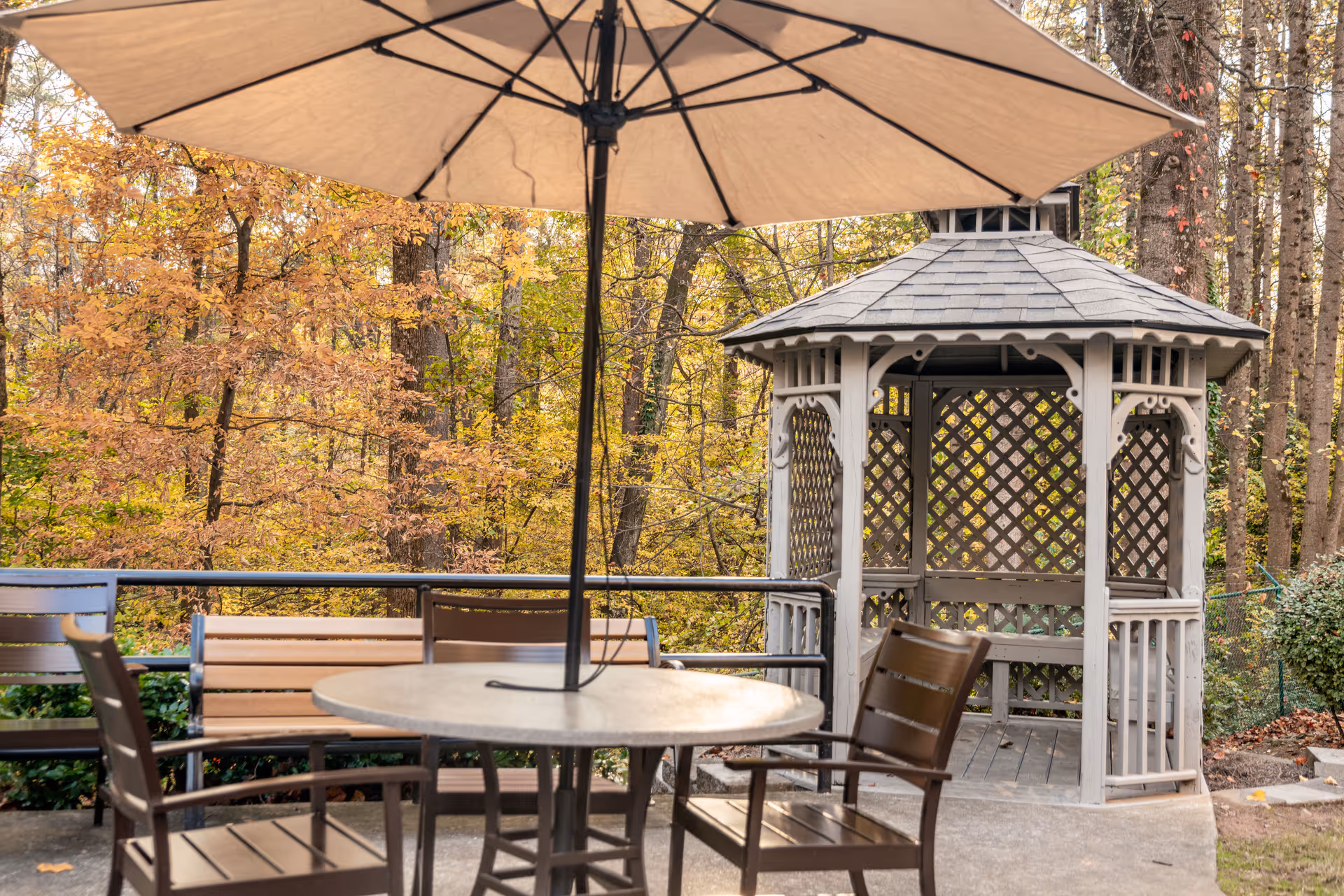 Outdoor patio area with a round table, chairs, and a large beige umbrella. In the background, there is a white wooden gazebo with lattice sides surrounded by trees with autumn-colored leaves.