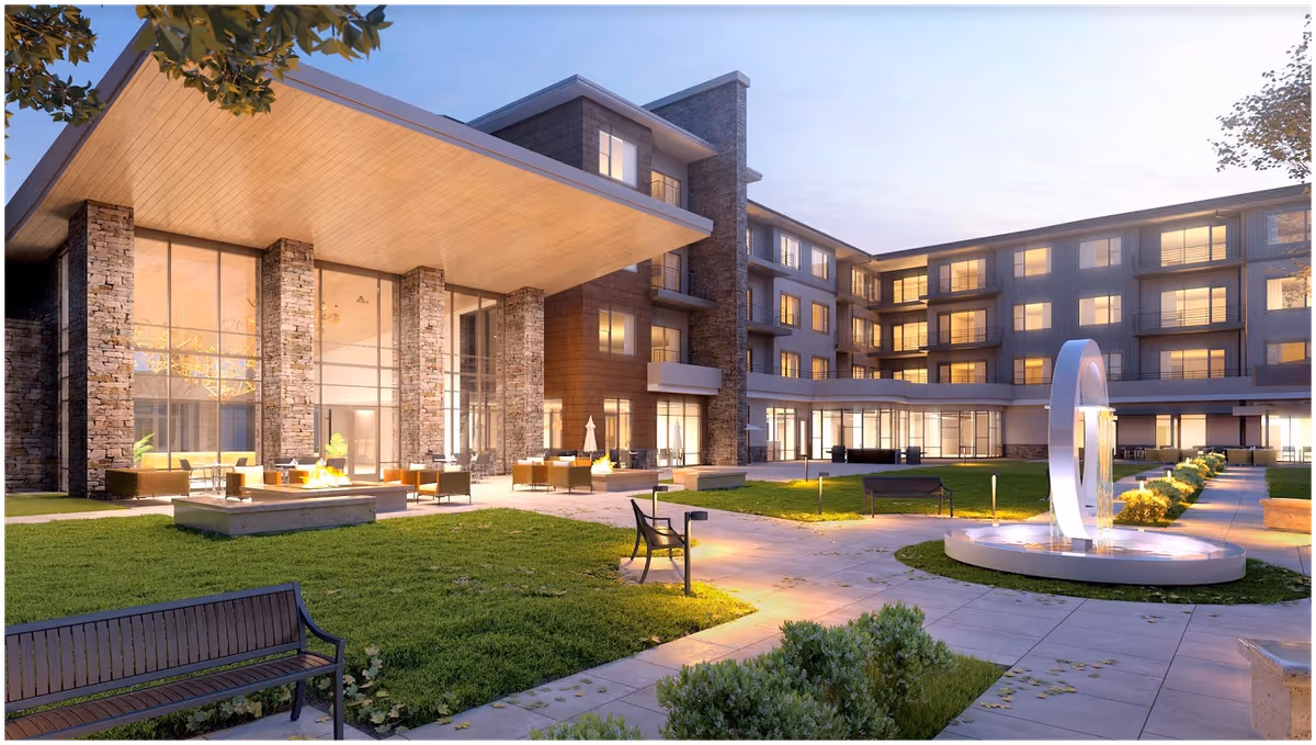 Outdoor courtyard area of The Ridge Pinehurst facility at dusk, featuring a modern building with large windows and stone pillars, outdoor seating with fire pits, benches, a circular water fountain, and well-maintained green lawns and pathways.