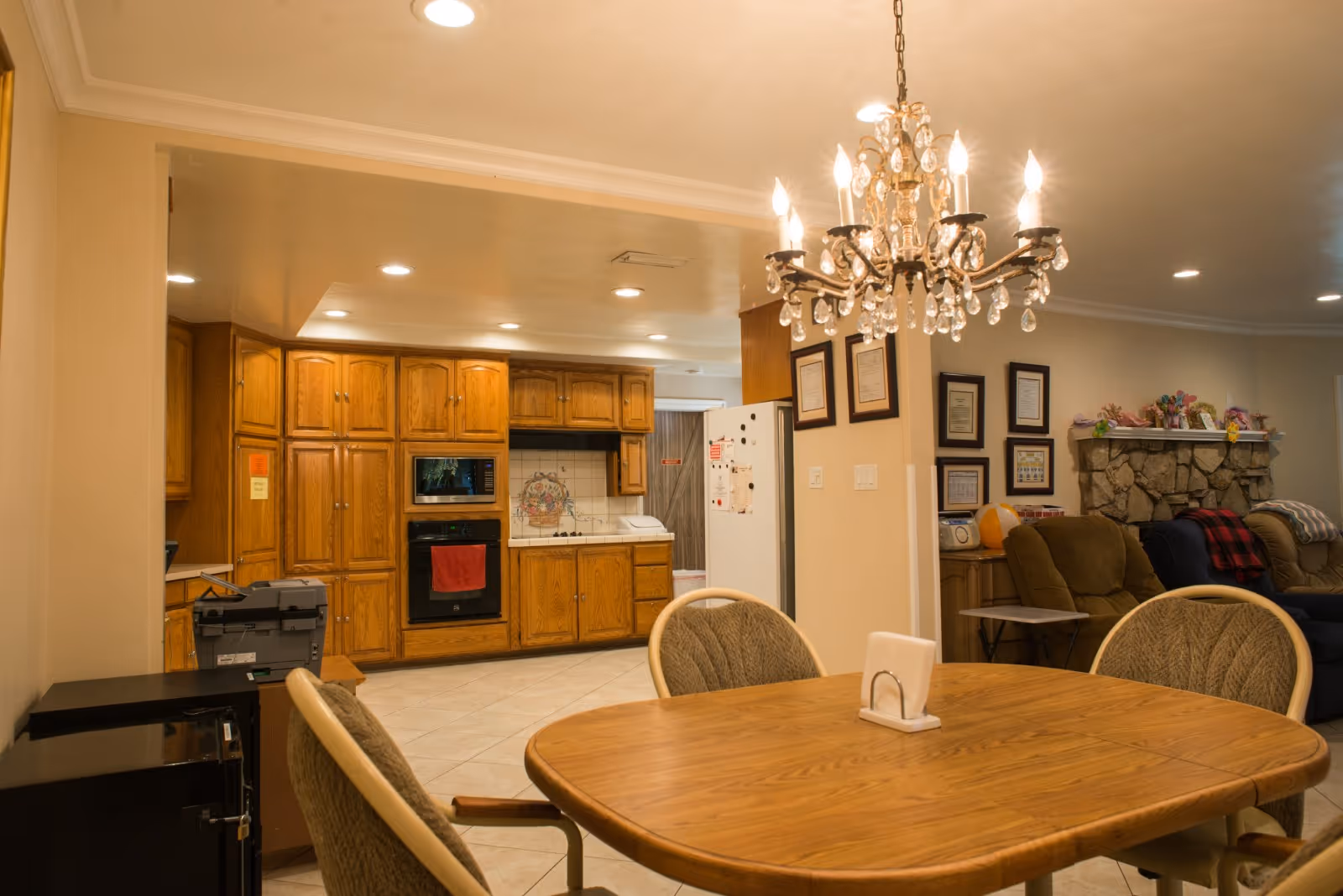 Interior view of a senior living facility showing a wooden dining table with four cushioned chairs, a chandelier hanging above, a kitchen area with wooden cabinets, an oven, microwave, and refrigerator, and a living room area with armchairs and a stone fireplace in the background.