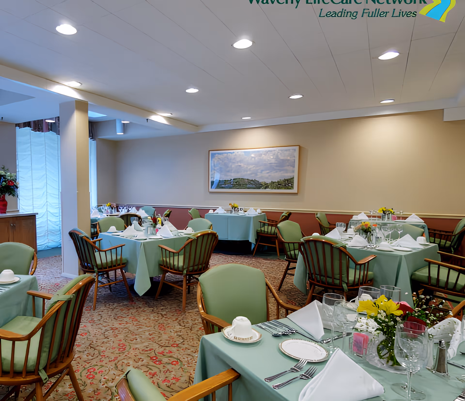 A dining room with multiple tables covered in light green tablecloths, each set with white napkins, plates, silverware, and glassware. The chairs have wooden frames with green cushions. There are floral centerpieces on the tables, a carpeted floor with a patterned design, and a framed landscape painting on the beige wall. The ceiling has recessed lighting.