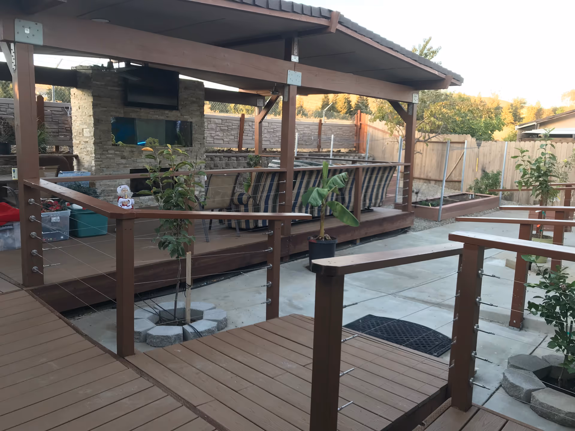 Outdoor patio area with wooden decking and railings, featuring a covered seating area with cushioned chairs and a stone fireplace with a mounted TV. There are small plants and trees in pots and planted in the ground, surrounded by a wooden fence and a stone wall in the background.