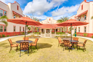 Outdoor courtyard area with green grass, three round tables each with four chairs, and red umbrellas providing shade. In the center background, there is a white gazebo. The courtyard is surrounded by a two-story building with white walls and red trim under a partly cloudy blue sky.