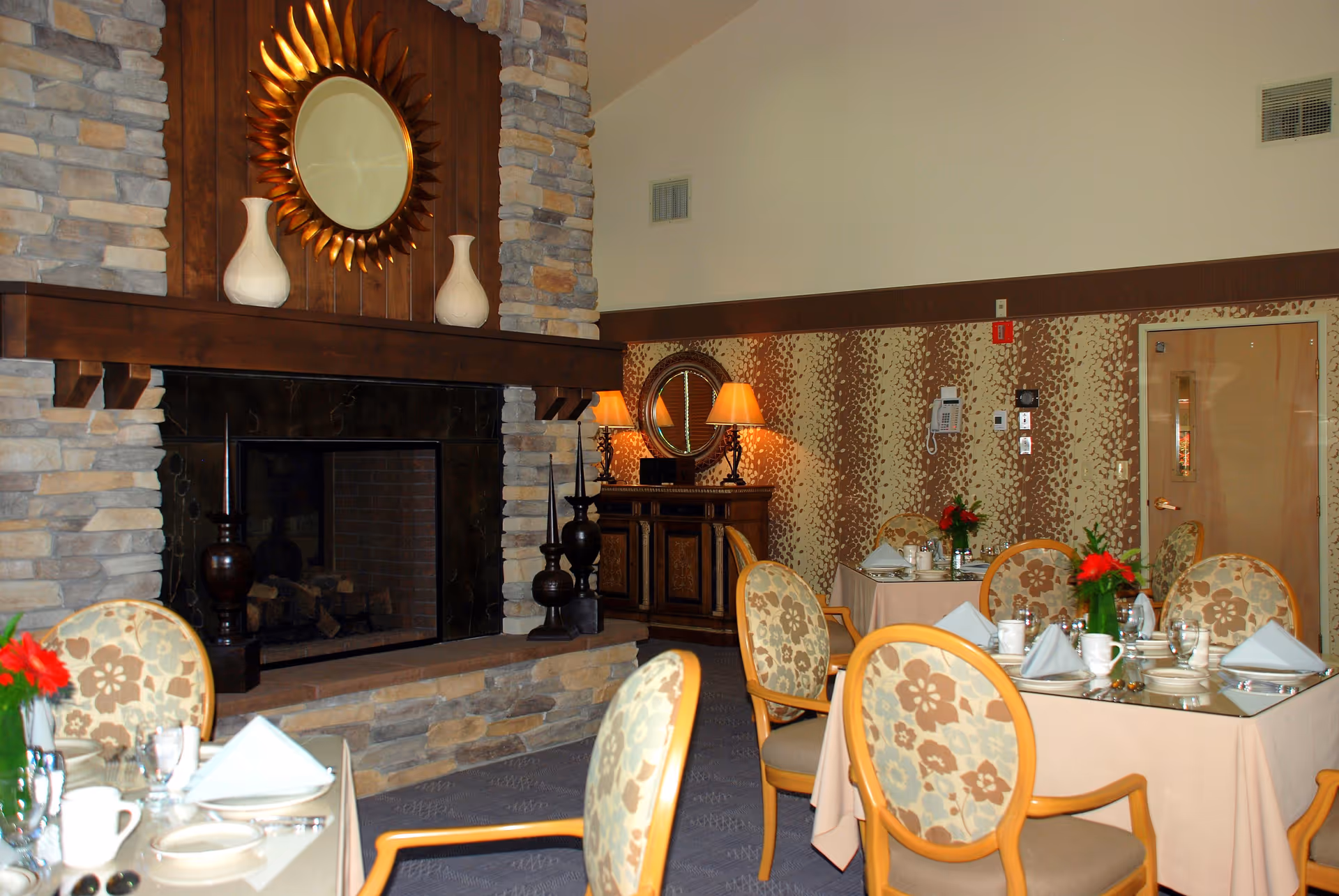 A cozy dining room with tables set for a meal, featuring floral-patterned chairs, beige tablecloths, and neatly folded napkins. The room has a stone fireplace with a wooden mantle, decorated with two white vases and a round sunburst mirror above it. In the background, there is a wooden sideboard with two lit table lamps and a round mirror on the wall.