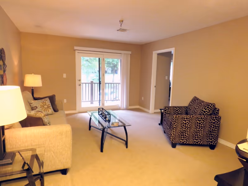 A cozy living room with beige walls and carpet, featuring a beige sofa with decorative pillows on the left, a glass coffee table in the center, and a patterned armchair on the right. There is a sliding glass door at the back leading to a balcony with a view of trees outside. A lamp is on a side table next to the sofa.