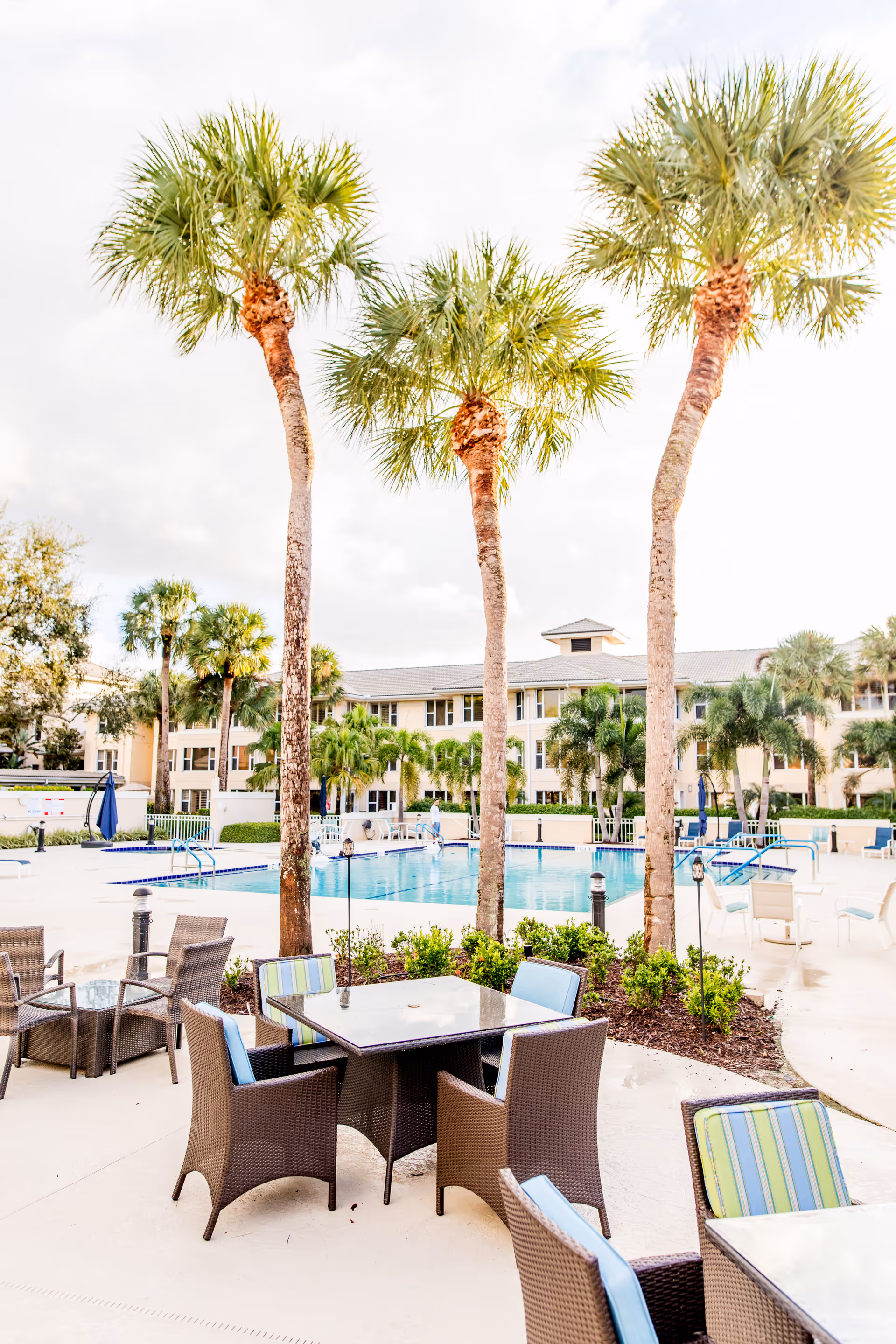 Outdoor seating area with wicker chairs and tables near a swimming pool surrounded by palm trees and a multi-story building in the background under a cloudy sky.