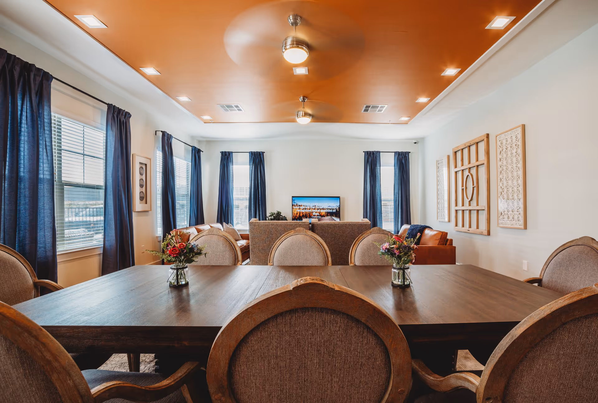 Dining room with a large wooden table and upholstered chairs facing a living area with a TV, blue curtains, and decorative ceiling lighting.