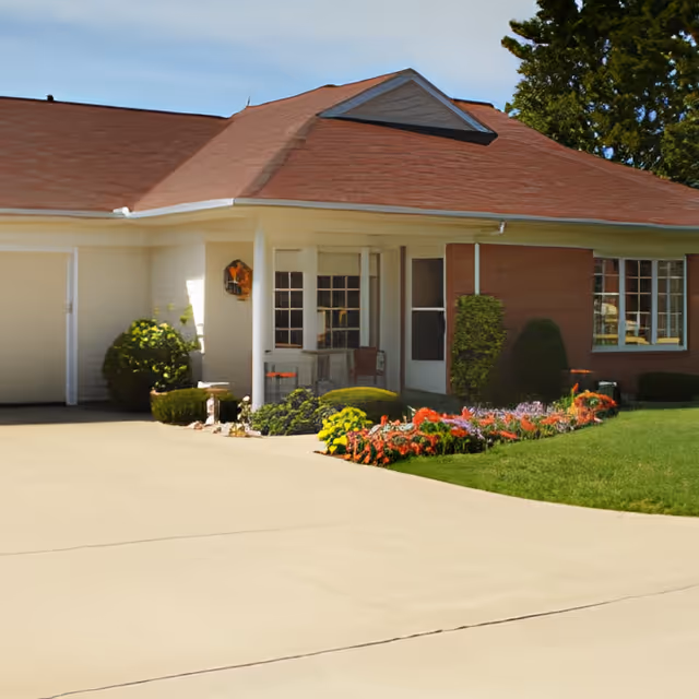 Front entrance of a single-story retirement community building with a covered porch, garage, driveway, and colorful flower beds.