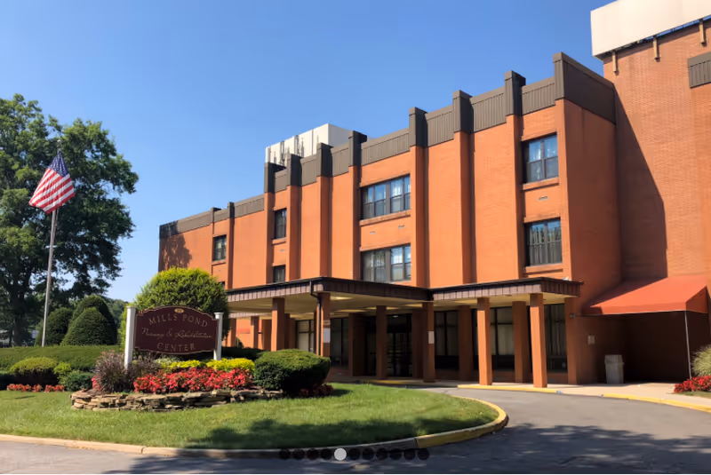 Exterior view of Luxor at Mills Pond Rehabilitation and Nursing Center with entrance and American flag.