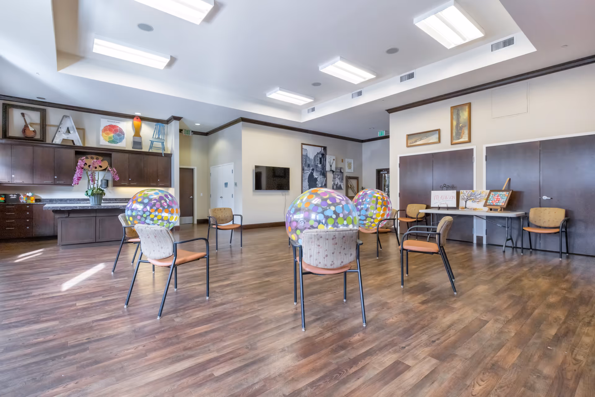 A spacious room with wooden flooring and several chairs arranged in a circle, each with a large colorful polka-dotted exercise ball placed on the seat. The room has bright overhead lighting, dark wood cabinets along one wall with decorative items on top, and framed artwork on the walls. There is a table with more chairs and art pieces on the right side of the room.