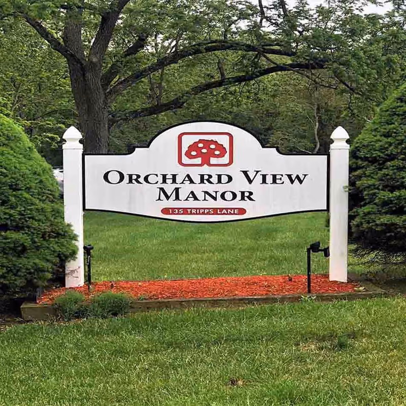 A white sign with black and red lettering that reads 'Orchard View Manor' with the address '135 Tripps Lane' below it, surrounded by green grass, trees, and bushes.