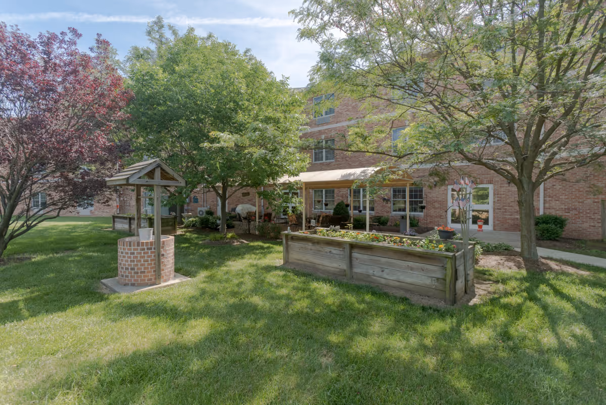 Sunlit courtyard with trees, a decorative brick well, raised wooden planters and a brick senior living building in the background.