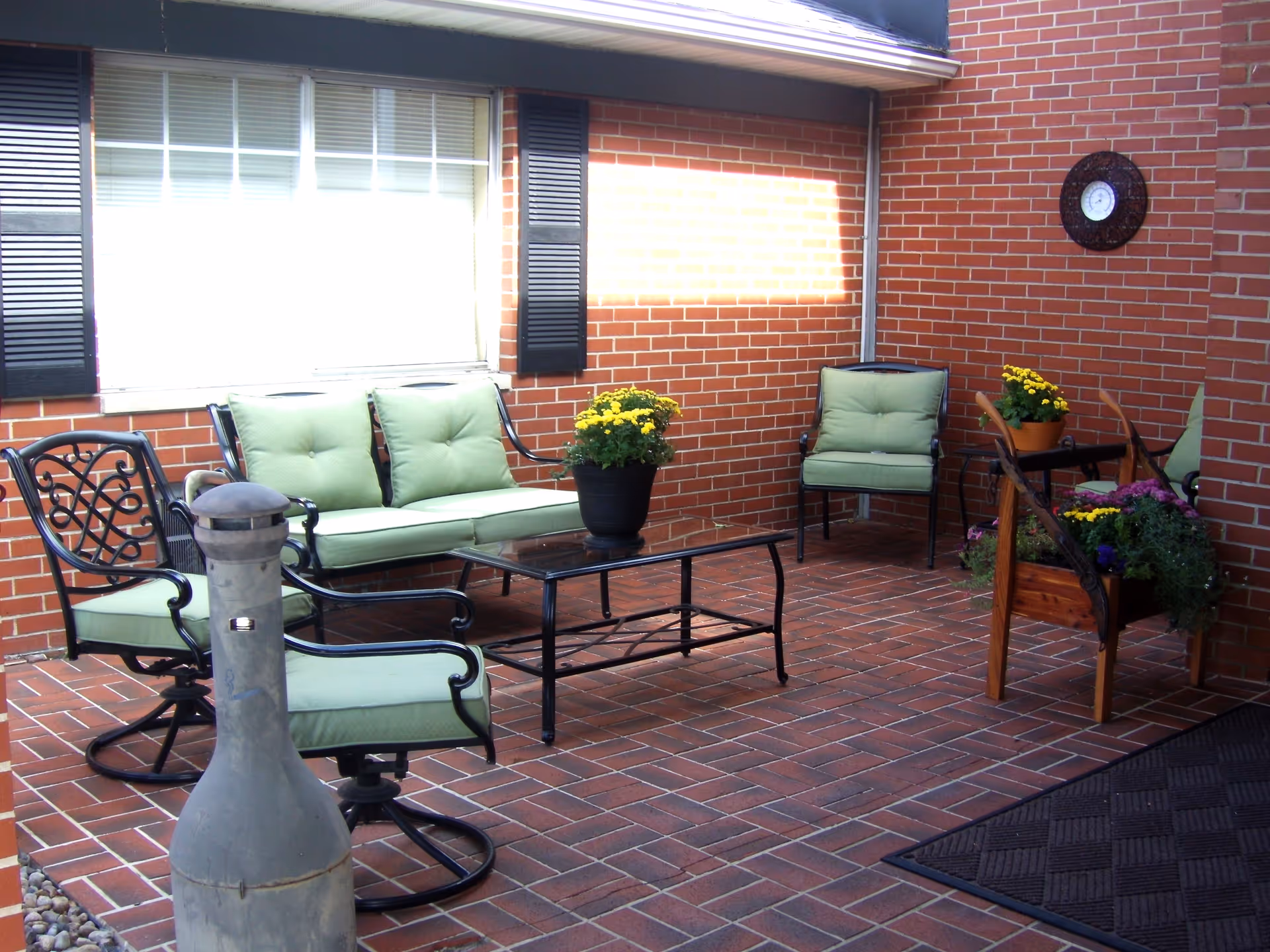A cozy outdoor patio area with brick walls and tiled floor, featuring green cushioned metal chairs and a loveseat around a glass-top coffee table. There are potted yellow flowers on the table and a wooden planter with various flowers. A decorative wall clock is mounted on the brick wall.