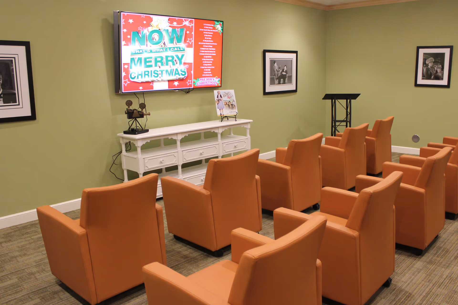 A small theater or media room with two rows of orange armchairs facing a wall-mounted flat screen TV displaying a Christmas-themed music playlist. The room has light green walls with two black and white framed photographs and a white console table beneath the TV holding decorative items.