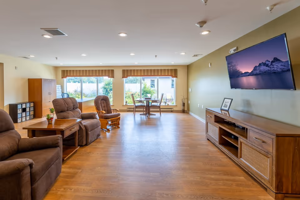 A spacious living room with wooden flooring featuring two brown recliner chairs and a wooden rocking chair around a wooden side table with a small plant. Large windows with striped valances allow natural light to fill the room. A flat-screen TV is mounted on a green accent wall above a wooden media console with a framed picture on it. In the background, there is a round table with four chairs near the windows.