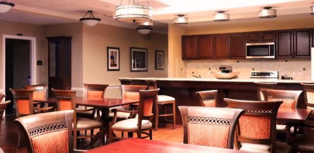 Interior view of a dining area with multiple wooden tables and chairs arranged neatly. In the background, there is a kitchen area with wooden cabinets, a microwave, stove, and a countertop with bar stools. The room is warmly lit with ceiling lights and decorated with framed pictures on the wall.