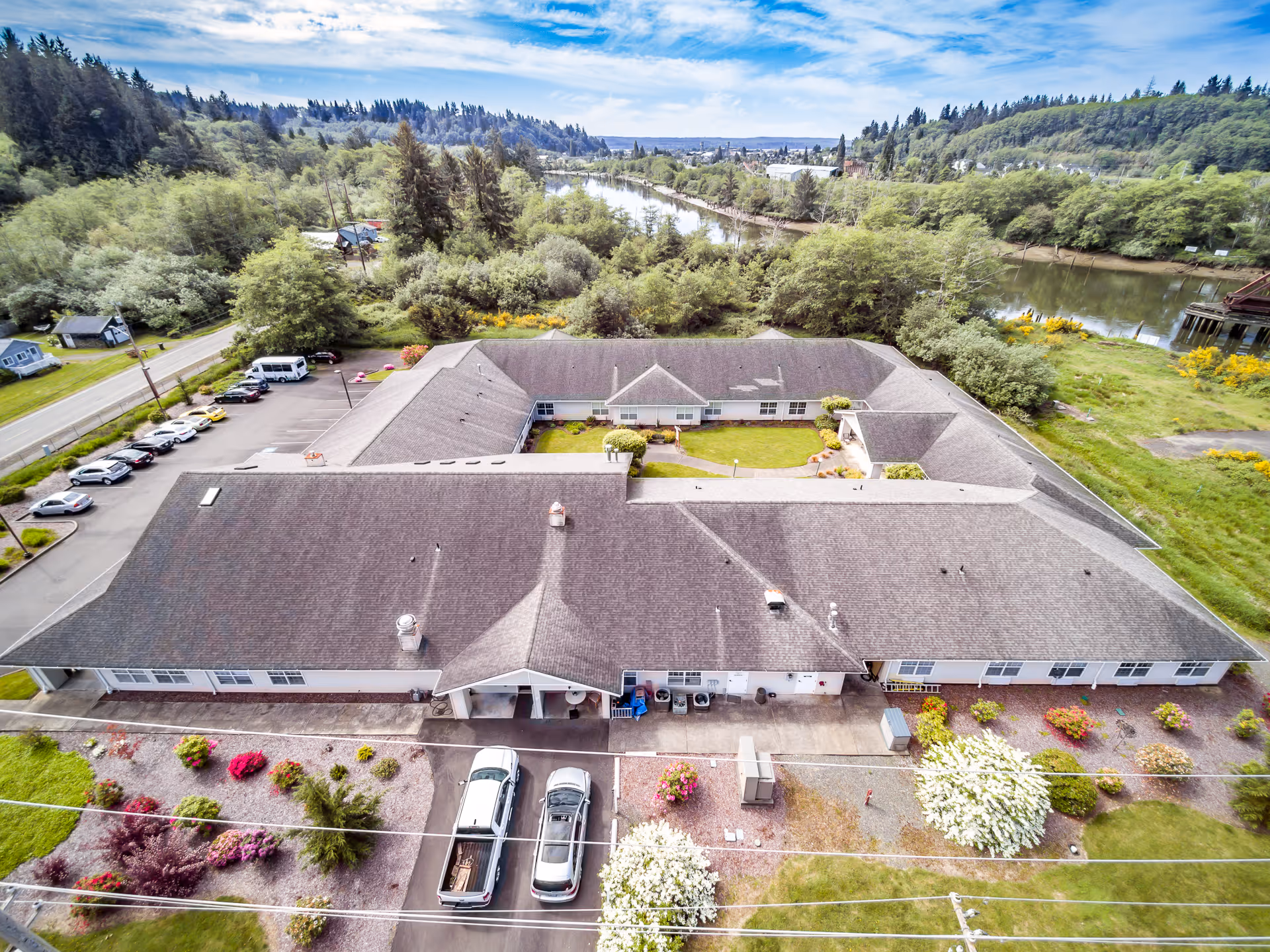 Aerial view of Riverside Place Memory Care facility showing a large, single-story building with a central courtyard. The building is surrounded by landscaped gardens with colorful flowers and shrubs. There is a parking lot with several cars on one side, and a river and forested hills in the background under a partly cloudy sky.