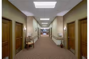 Carpeted interior corridor of a senior living facility with wooden doors on both sides, handrails, chairs, and overhead lighting.