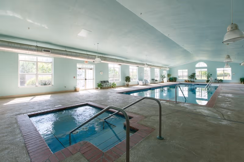 Indoor swimming pool area with a smaller hot tub in the foreground and a larger swimming pool in the background. The room has large windows letting in natural light, light blue walls, and several potted plants along the walls. There are handrails for both the hot tub and the pool, and ceiling lights hang from the high ceiling.